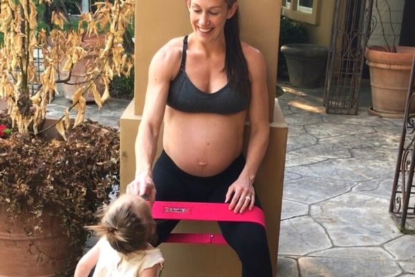 fit pregnant mom in a gray sports bra playing with her toddler and doing a wall squat with a pink loop band around her knees