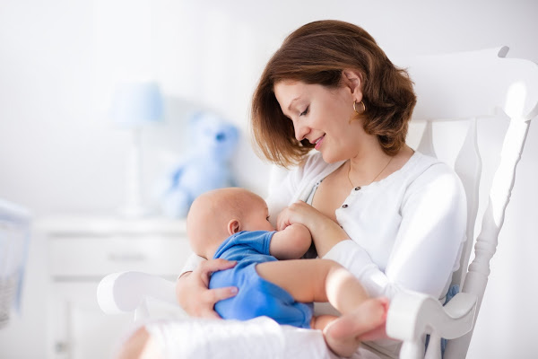 Young mother holding her newborn child. Mom nursing baby. Woman and new born boy in white bedroom with rocking chair and blue crib. Nursery interior. Mother playing with laughing kid. Family at home