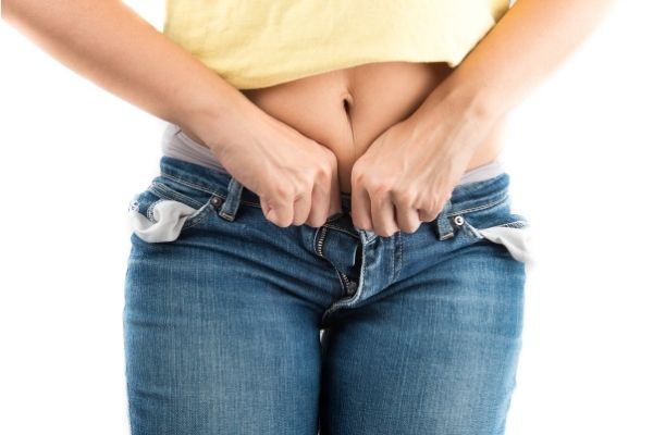 women wearing a yellow shirt with stomach showing trying to zip her jeans button with difficult from fat