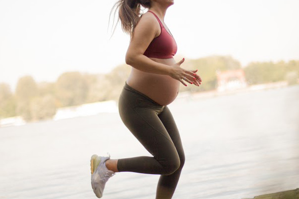 A beautiful pregnant woman running on the nature trail  She is in her third trimester and wearing red maternity sports top and leggings. She's been a runner and continues to jog regularly to stay fit and strong during pregnancy. Being physically active is important for a healthy pregnancy.
