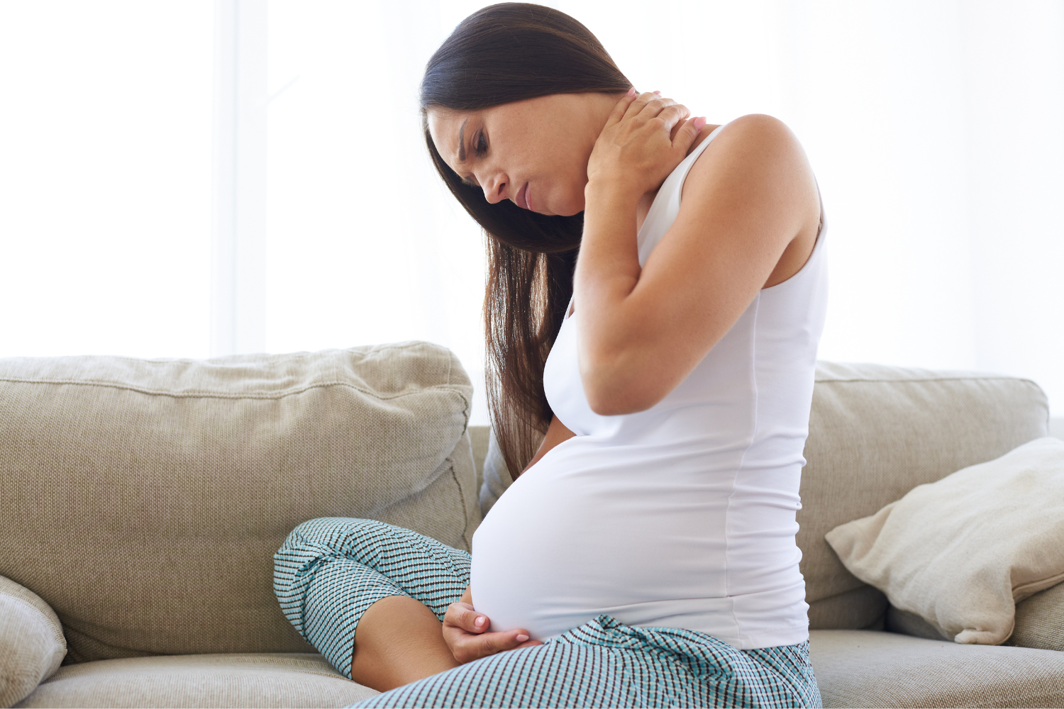 pregnant mom in a white tanktop and pajama pants sitting on a couch holding her neck with a look of pain on her face.