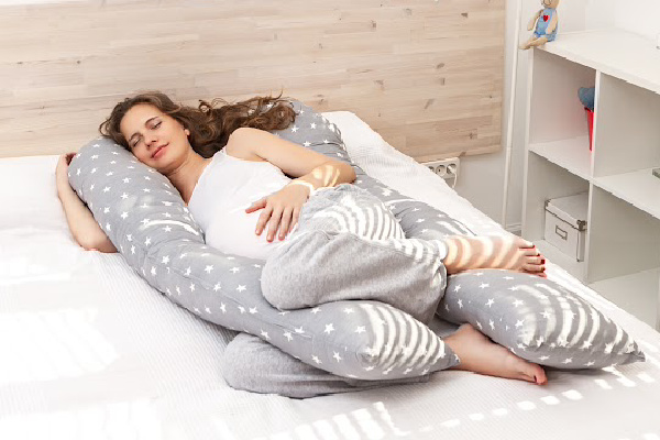 tired pregnant mom wearing a white tank top sleeping with a body pillow that supports he belly and back to improve sleep