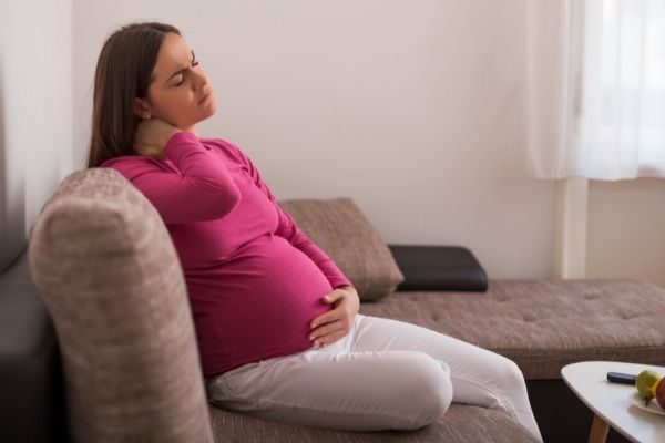 pregnant mom in a pick top and white pants sitting on the couch holding her neck in pain