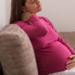 pregnant mom in a pink shirt and white pants sitting on a couch hold her neck in pain