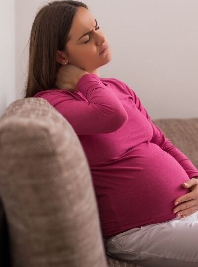 pregnant mom in a pink shirt and white pants sitting on a couch hold her neck in pain