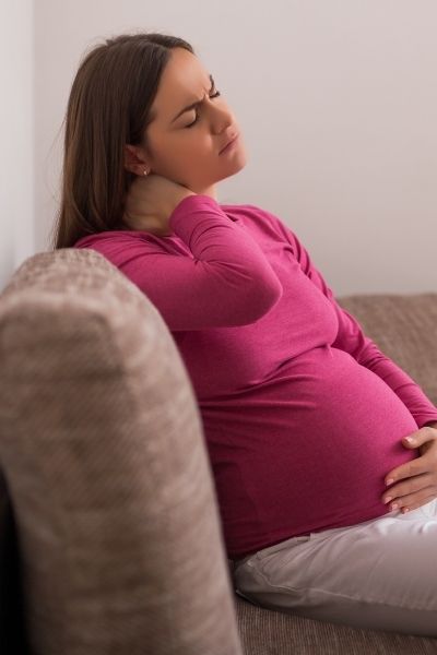pregnant mom in a pink shirt and white pants sitting on a couch hold her neck in pain