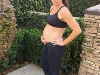 Fit pregnant mom in black pants and black sports bra holding her pregnant belly in the second trimester