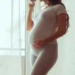 pregnancy mom standing in front of the window with hand on her belly, drinking coffee